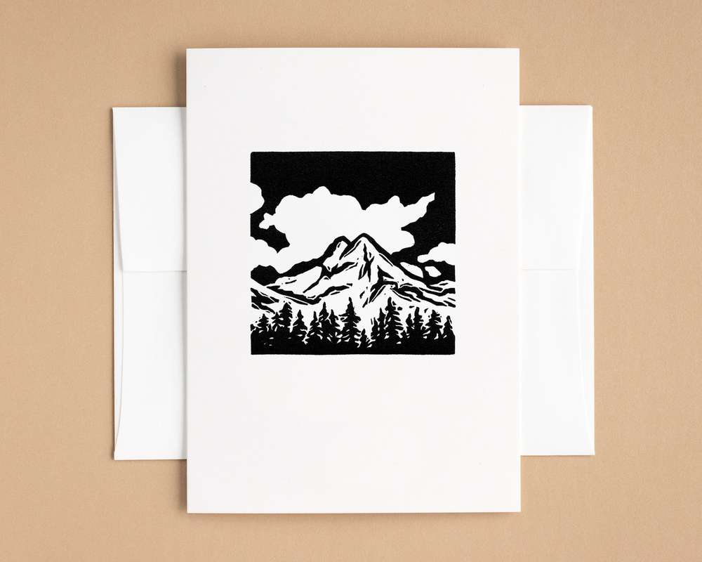 A vertical white card with a black and white depiction of a mountain peak and trees. The card sits on top of a white envelope, which lies on top of a brown backdrop.